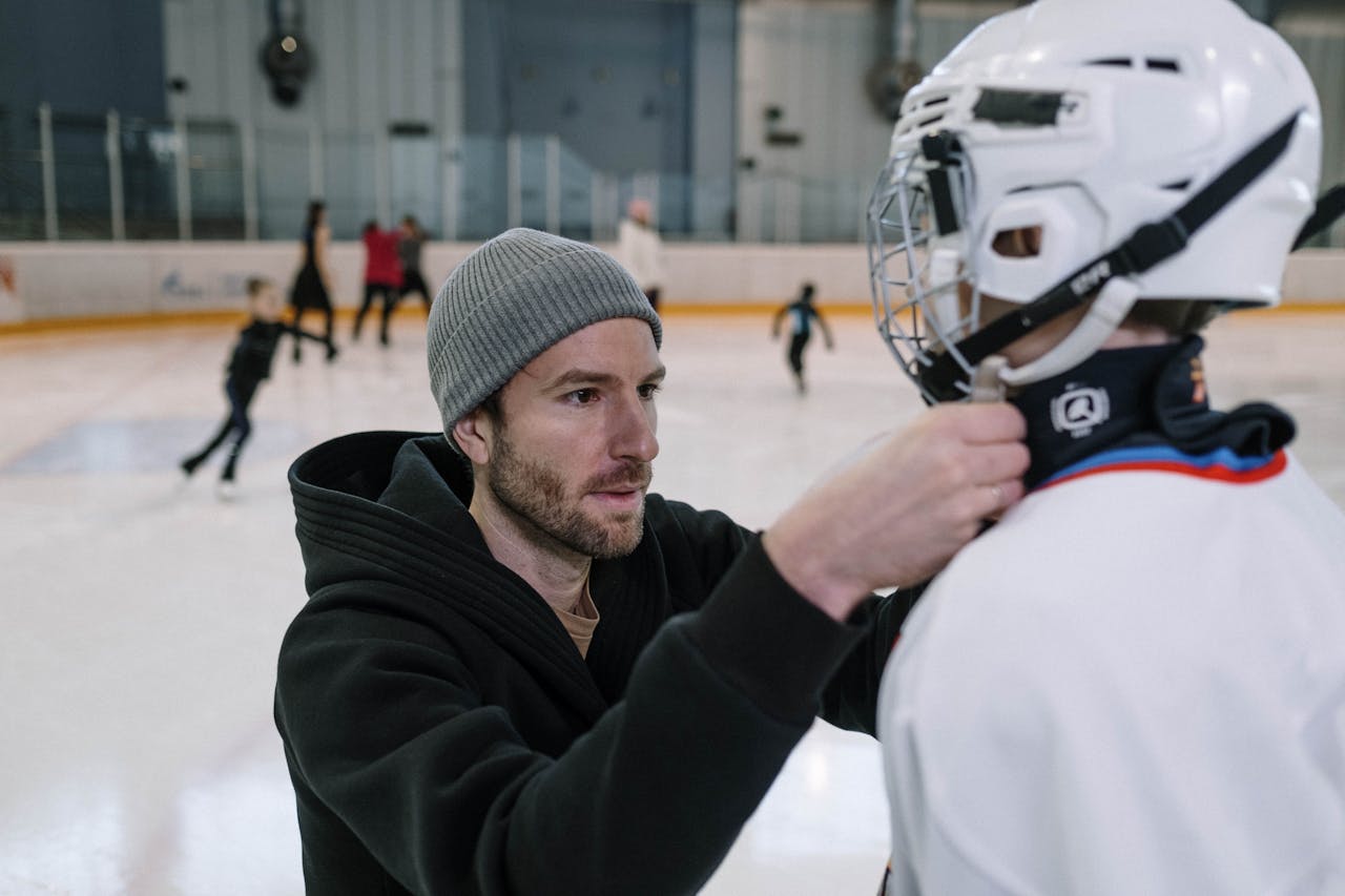 Father helps adjust sons hockey gear at an indoor ice rink, pre-game preparation.